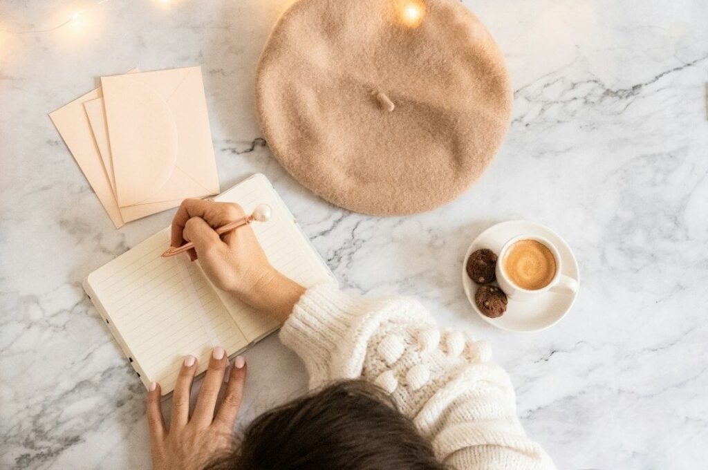 Person skriver i en anteckningsbok med kaffe och basker på bordet